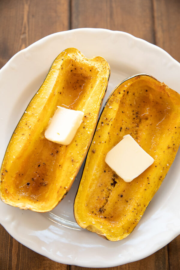 Two halves of a delicata squash sit on a white plate. They have been roasted with maple syrup and black pepper. Pats of butter are melting into the squash.