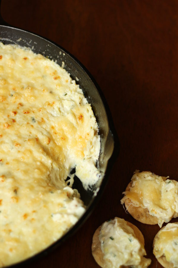 A cast iron skillet is on a wooden table. The skillet is full of artichoke parmesan dip - cheesy, gooey, and nicely browned on top. A few crackers topped with dip sit nearby.