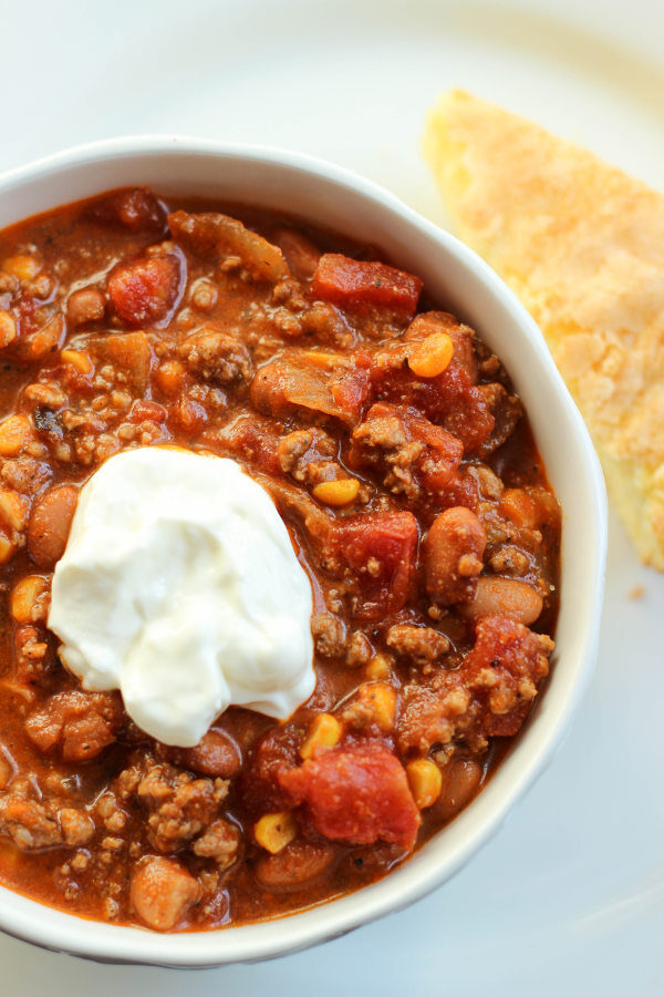 A big white bowl is full of meaty beef and bean chili. You can see chunks of tomatoes and pieces of corn. There is a dollop of sour cream on top. 
