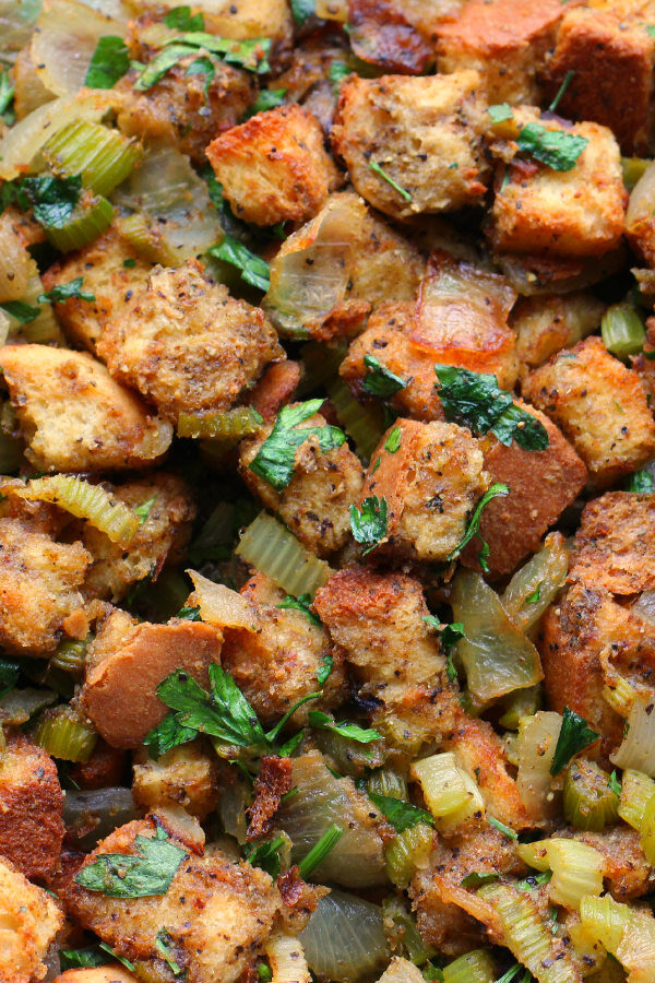 An up close image of turkey stuffing. You see big chunks of golden bread, onions, celery, and plenty of bright herbs.