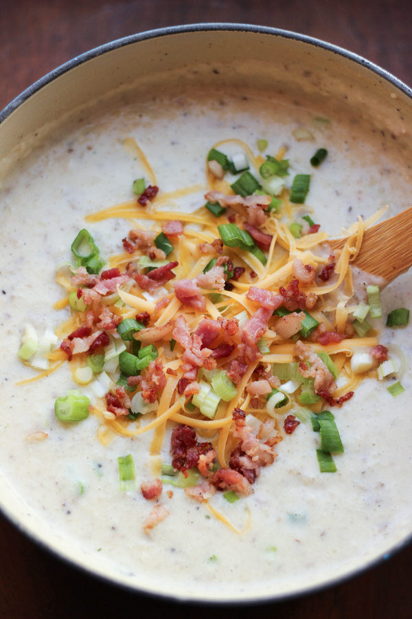 A big pot of loaded baked potato soup sits ready to eat with a wooden serving spoon in it. You can see piles of crumbled bacon, green onions, and cheddar cheese on top. 