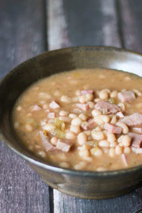 Brown pottery bowl full of ham and bean soup. You see a rich broth, white navy beans and big chunks of ham in the bowl.