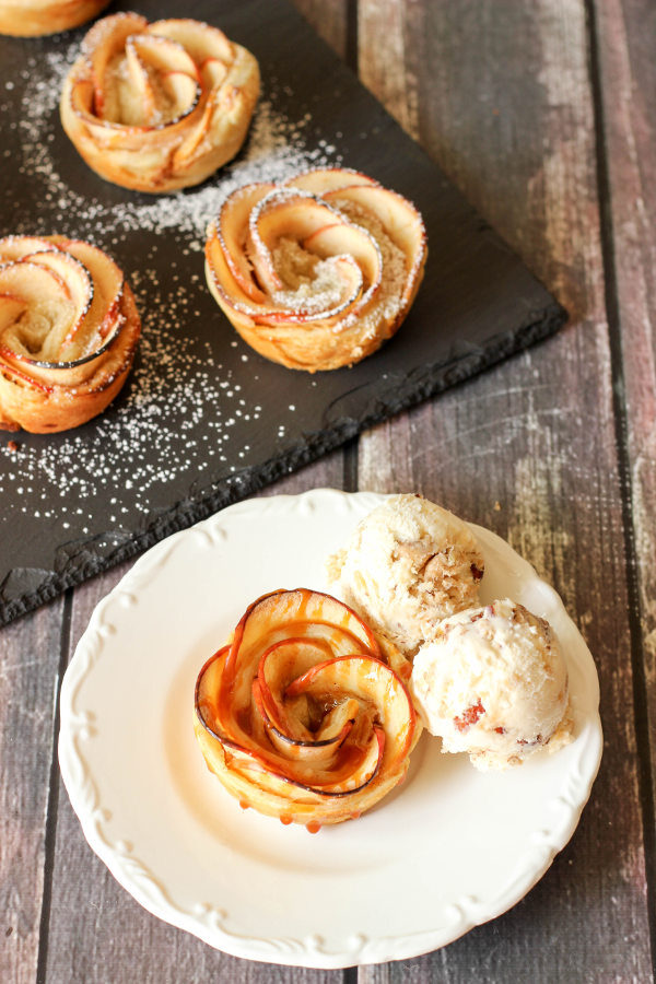 A beautiful apple pie rose sits on a white plate. There are scoops of ice cream on the side and a bit of caramel sauce drizzled over the top of the rose.