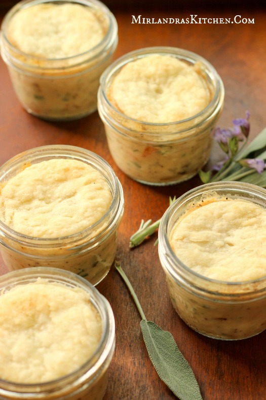 Four individual chicken pot pies are baked in mason jars and sitting on a wooden table.  Some fresh herbs are nearby.