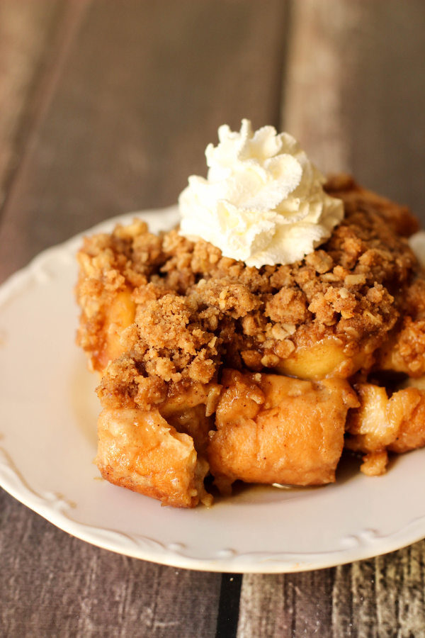 A big square of apple crisp French Toast casserole sits on a white plate. You can see a dollop of whipped cream on top.