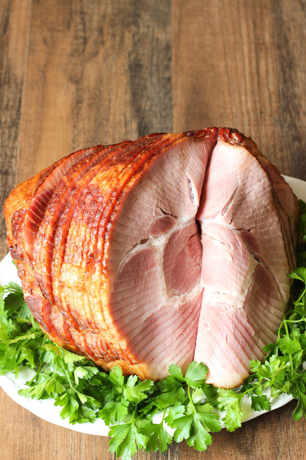 A large spiral ham is sitting on a bed of greenery and white platter waiting to be served for the holidays. The ham is glazed with a lovely brown sugar honey glaze.