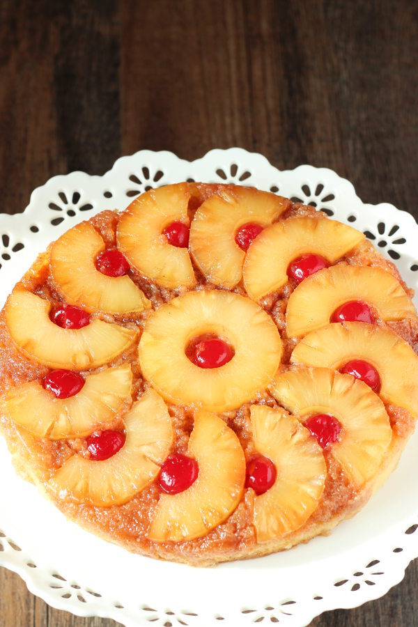 A beautiful pineapple upside down cake is displayed on a white cake stand. The pineapple is cut into half circles and arranged like a pinwheel around a center ring of pineapple. Cherries sit inside each ring or half ring.