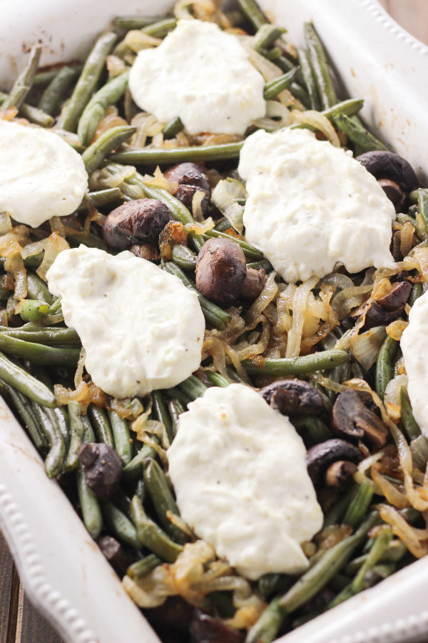 A big rectangular white pan is full of green beans. You can see lots of caramelized onions and roasted mushrooms in this gluten free green bean casserole. There are also big pillows of cheesy, creamy sauce dolloped on top.