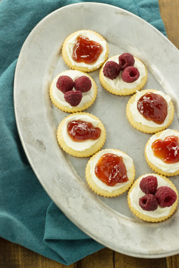 A silver oval platter has crackers on it. The crackers have cream cheese, jam, and raspberries as toppings.