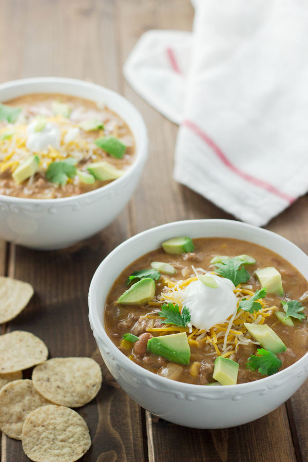 Two bowls of cheese taco soup sit on a wooden table .Each bowl is garnished with cheese, cilantro, sour cream and chunks of avocado.