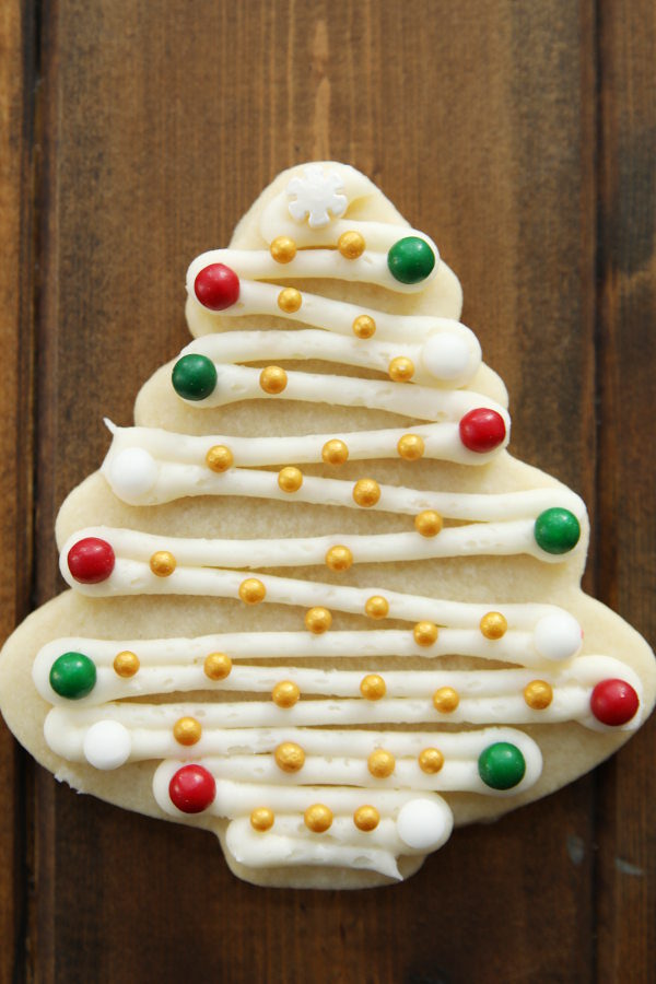A cut out cookie in a Christmas tree shape is decorated with white buttercream and green, red, white and gold balls like ornaments.