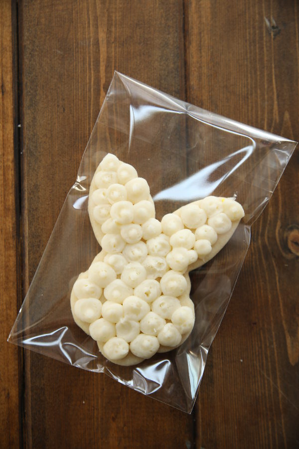 A bunny face and ears cut out sugar cookie is decorated with swirls of white frosting. It is packaged in a self sealing plastic bag for gifting.