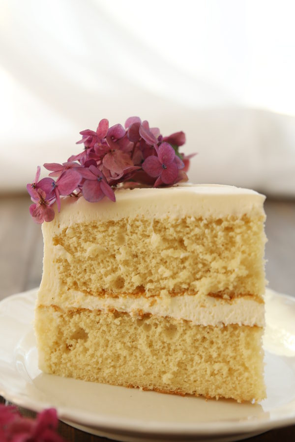 A big wedge of vanilla cake sits on a white plate. Both layers are stacked together with buttercream frosting and some purple hydrangea flowers decorate the cake on top. 