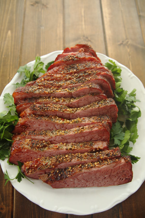 A slab of oven roasted corned beef is sliced thickly on a white plate. There is green, leafy herbs for garnish on the sides.