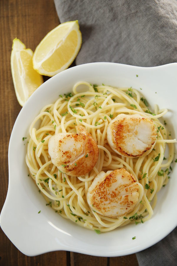 A round white dish holds buttery pasta topped with three large golden crusted scallops. There are wedges of lemon and a gray napkin nearby.