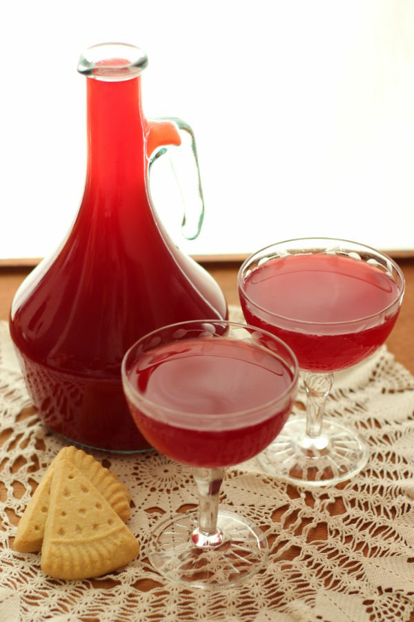 A jug of raspberry cordial from the Anne of Green Gables Story sits with some shortbread biscuits and a few classes of cordial on a lace doily.