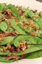 tender green snow peas are sauteed with bacon and topped with crispy french fried onions on a white plate.