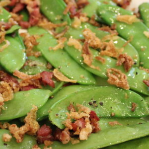 tender green snow peas are sauteed with bacon and topped with crispy french fried onions on a white plate.