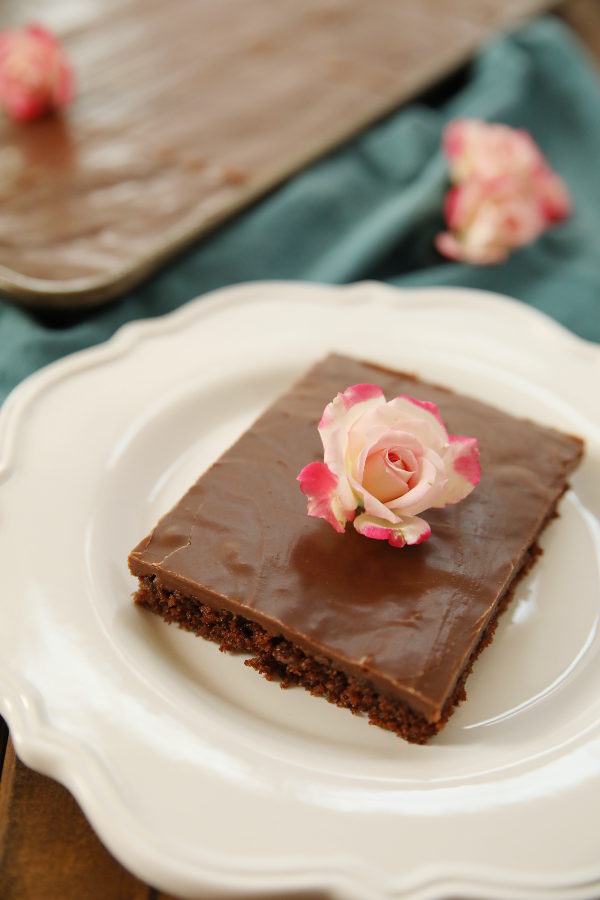A big slice of Texas sheet cake sits on a white plate. The pan of cake is in the background and a few pink roses are scattered around.