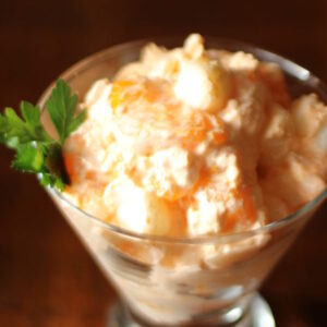 An elegant stemmed glass is full of orange fluff salad. You can see mini marshmallows and orange slices in the salad. It is garnished with grenery.