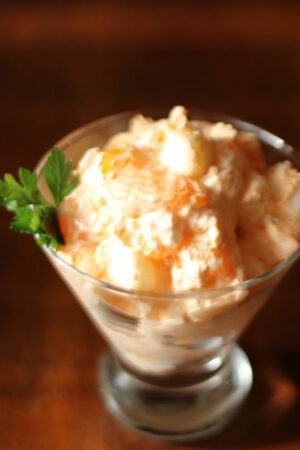 An elegant stemmed glass is full of orange fluff salad. You can see mini marshmallows and orange slices in the salad. It is garnished with grenery.