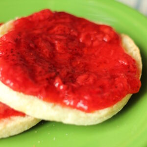strawberry freezer jam is slathered in a thick layer on toasted English muffins. Two muffins with jam rest on a bright green plate.