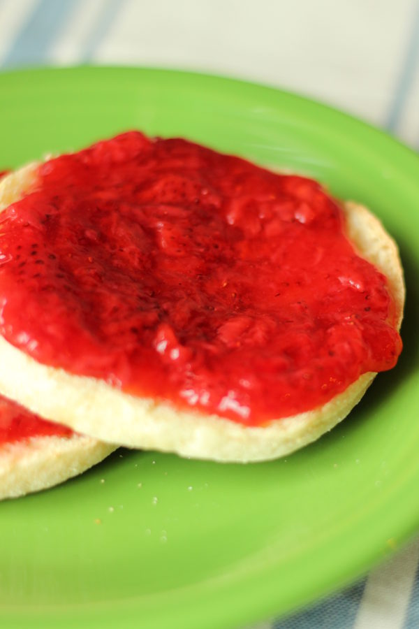 strawberry freezer jam is slathered in a thick layer on toasted English muffins. Two muffins with jam rest on a bright green plate.