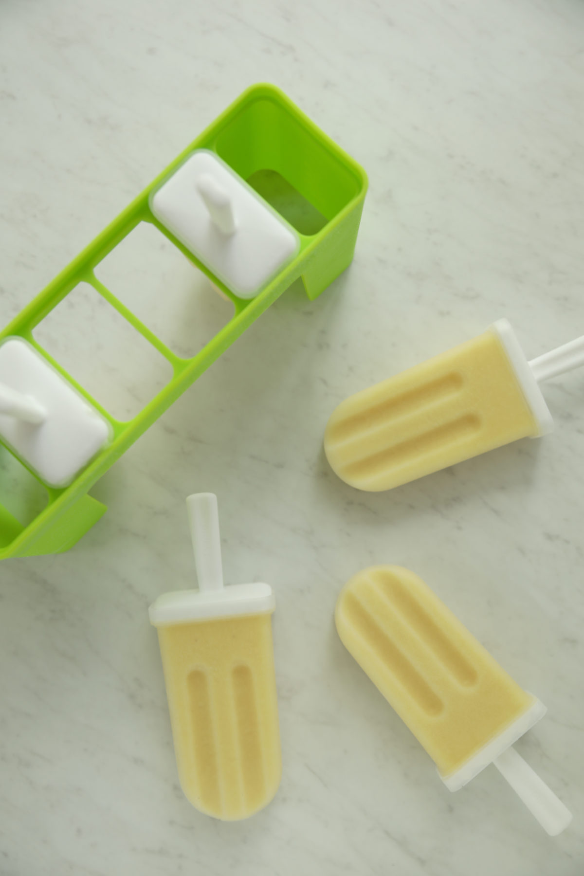 Piña colada popsicles unmolded next to a green silicone popsicle mold on a marble surface.