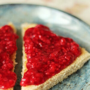 A slice of toast is cut into two triangles and slathered with no cook raspberry jam. The toast is on a blue stoneware plate.