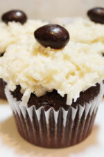 Three almond joy cupcakes sit on a white plate. You can see the chocolate cupcake, the coconut frosting and a chocolate coconut almond on top.