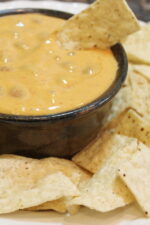 A pottery bowl full of chili con queso sits surrounded by chips. There is a chip stuck into the dip.