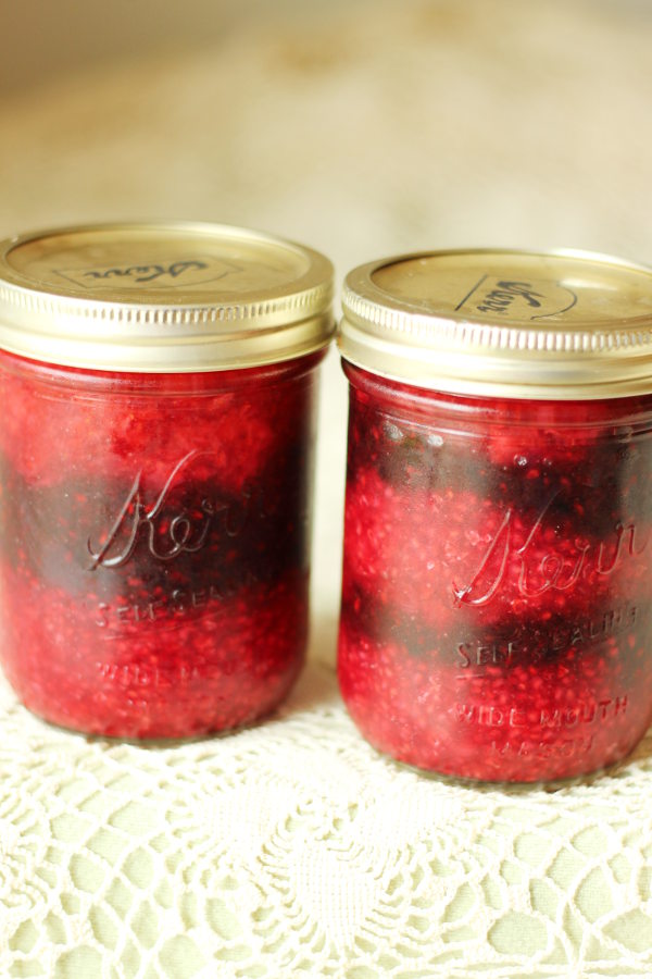 Two jars of no cook berry jam. The jam has been layered into the jar in stripes so you can see a layer of raspberry, layer of blackberry, and layer of strawberry.