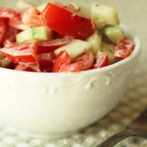 A white bowl is full of creamy tomato and cucumber salad with a mayonnaise dressing. The bowl is sitting on a green and white checked table cloth next to an antique silver fork.