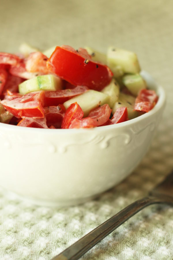 A white bowl is full of creamy tomato and cucumber salad with a mayonnaise dressing. The bowl is sitting on a green and white checked table cloth next to an antique silver fork.