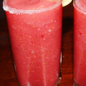 Two tall glasses are full of a frozen strawberry cocktail. You can see sugar on the rim and slices of lemon on the edge of the glass.