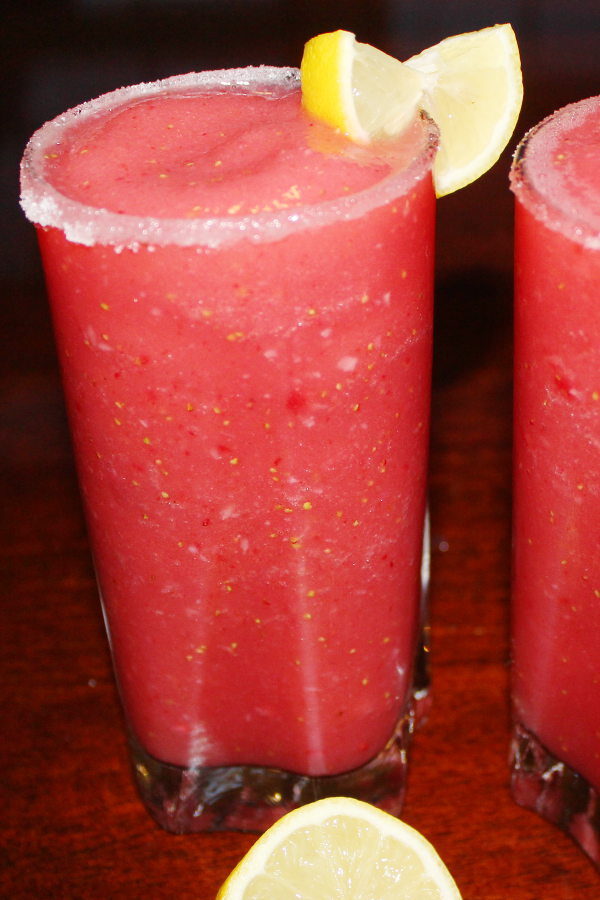 Two tall glasses are full of a frozen strawberry cocktail. You can see sugar on the rim and slices of lemon on the edge of the glass.