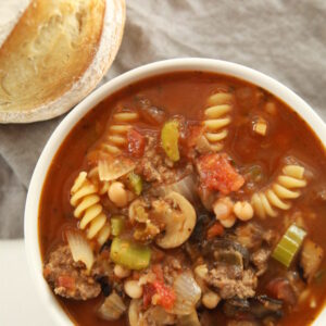 A hearty bowl of beef minestrone soup sits on a white table. There is a gray cloth on the rustic farmhouse table and a loaf of artisan bread next to the bowl. The soup is full of mushrooms, carrots, celery, and pasta.