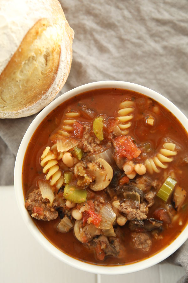 A hearty bowl of beef minestrone soup sits on a white table. There is a gray cloth on the rustic farmhouse table and a loaf of artisan bread next to the bowl. The soup is full of mushrooms, carrots, celery, and pasta.