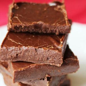 Five pieces of easy fudge are stacked on a white plate. The fudge is creamy and soft in the photo.