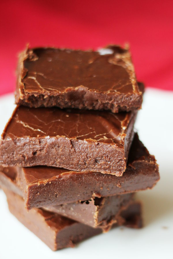 Five pieces of easy fudge are stacked on a white plate. The fudge is creamy and soft in the photo.