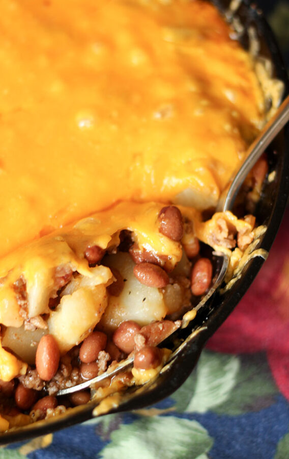 Poor Potato Hamburger Casserole is baked in a cast iron pan. The casserole is full of beans, potatoes, hamburger and delicious sauce. The mixture is topped with cheddar cheese.