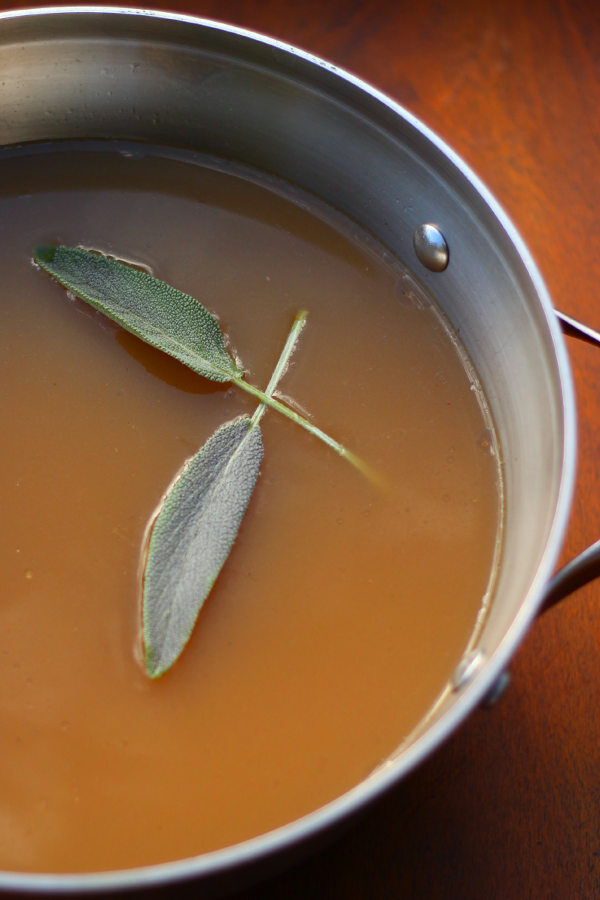 A round stainless steel pan is full of beef stock made from roasted beef bones. The stock is a dark amber color and garnished with sage leaves.