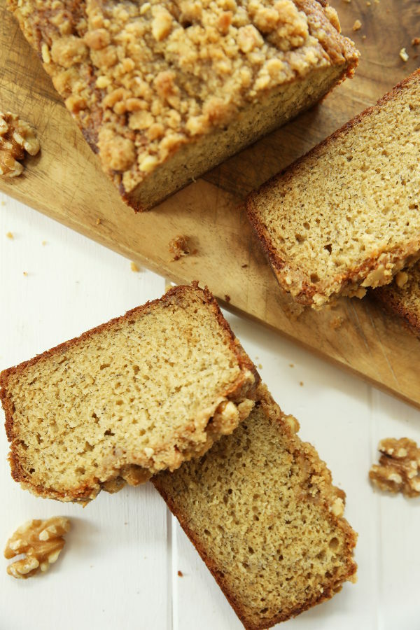 This easy banana bread is sitting on a cutting board. Several slices have been cut off and arranged on the cutting board and main table. There are several walnuts around.