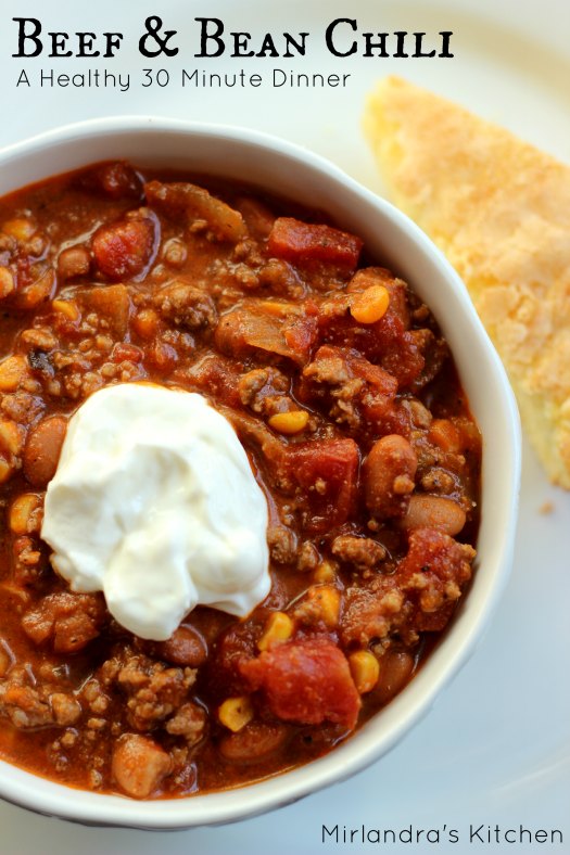 A big bowl of beef and bean chili. The chili is garnished with a dollop of sour cream and sitting next to a wedge of sweet corn bread.