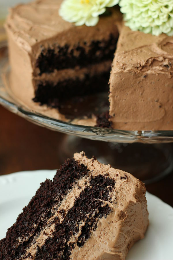 A beautiful black magic cake sits on a clear cake stand. There is one slice out of the cake on a white plate in front. The top of the cake has a few white zinnias on it. 