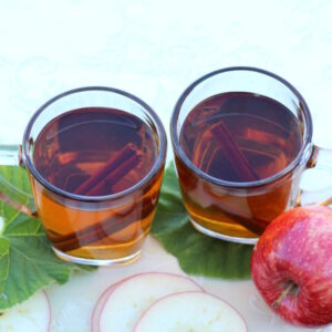 Two cups of mulled apple cider are ready to drink. Apple slices are nearby on the table.