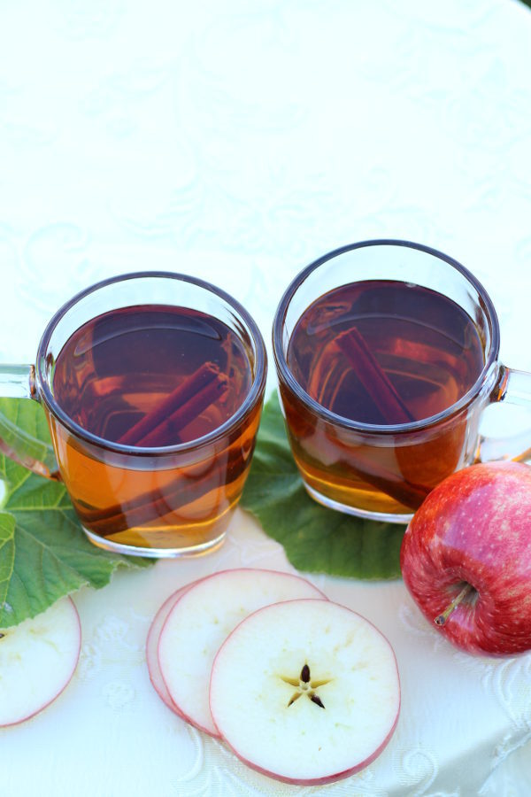 Two cups of mulled apple cider are ready to drink.  Apple slices are nearby on the table.