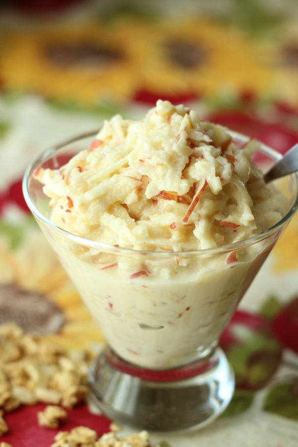 A clear glass bowl of apple salad sits on a table. There is granola scattered around and a spoon sticking out. You can see that the apple is grated and covered in a yogurt dressing.