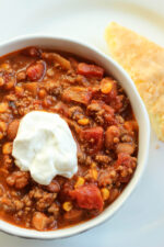 A big white bowl is full of meaty beef and bean chili. You can see chunks of tomatoes and pieces of corn. There is a dollop of sour cream on top.