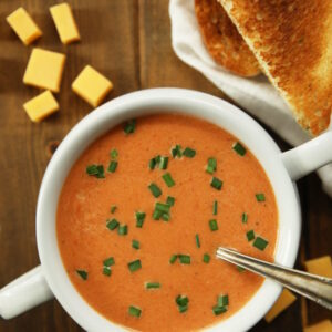 A white soup bowl with two handles is full of homemade tomato soup. The soup is a rich orange and garnished with some chopped chives. An old fashioned silver spoon is in the soup and chunks of cheddar cheese sit nearby as does a some toasted bread.